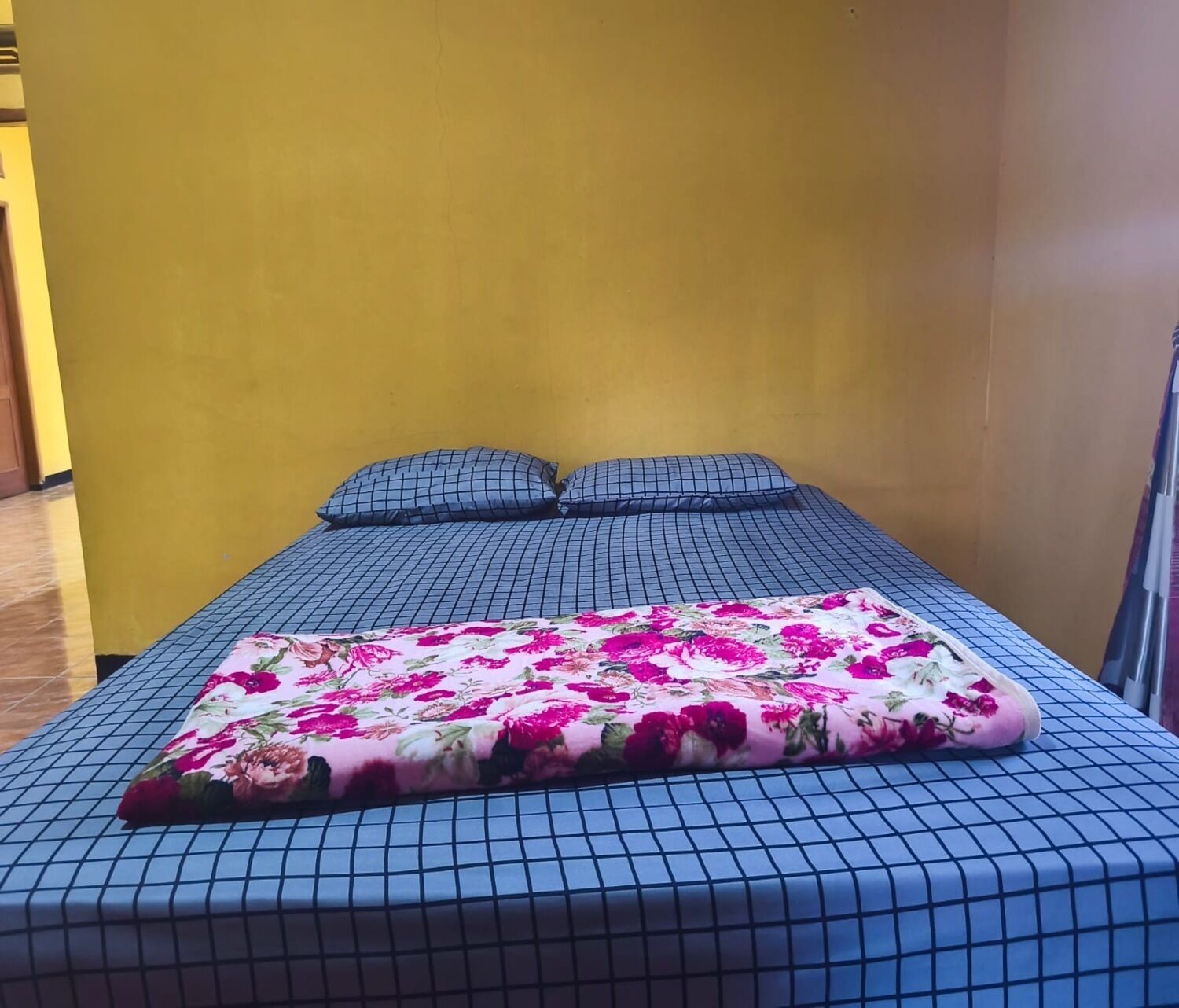 Una cama con colcha estampada de flores y otra colcha azul a cuadros, dos almohadas y una pared amarilla.