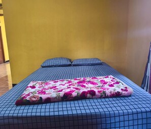 Una cama con colcha estampada de flores y otra colcha azul a cuadros, dos almohadas y una pared amarilla.