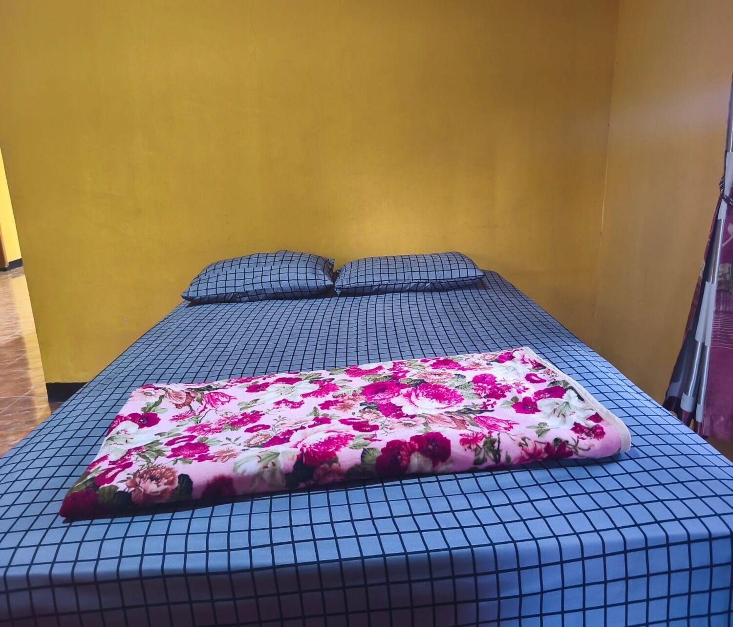 A bed with a blue checkered bedspread and a floral throw blanket, two matching pillows, and a yellow wall.