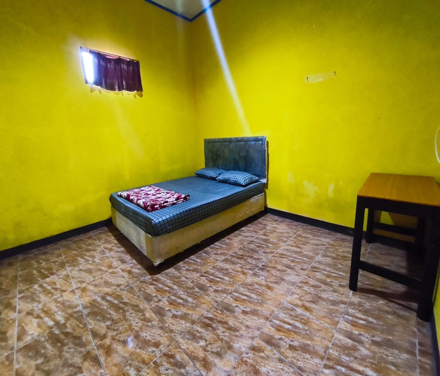 Una stanza piccola con un letto singolo, pavimento piastrellato, un tavolo di legno e una finestra con tenda.