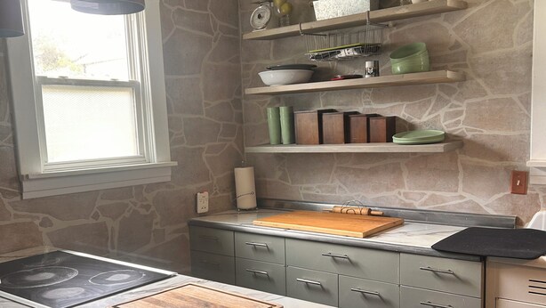 A kitchen with a stone backsplash, a marble countertop, a wooden cutting board, and a hanging pendant light.
