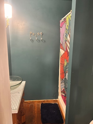 A bathroom with a blue wall, a glass sink, a wooden floor, a black rug, and a colorful patterned shower curtain.