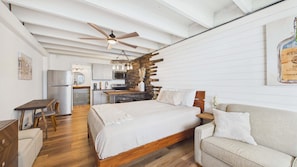 Ein Schlafzimmer mit Bett, Holz-Kopfteil, Deckenventilator und einer kleinen Küchenzeile im Hintergrund.