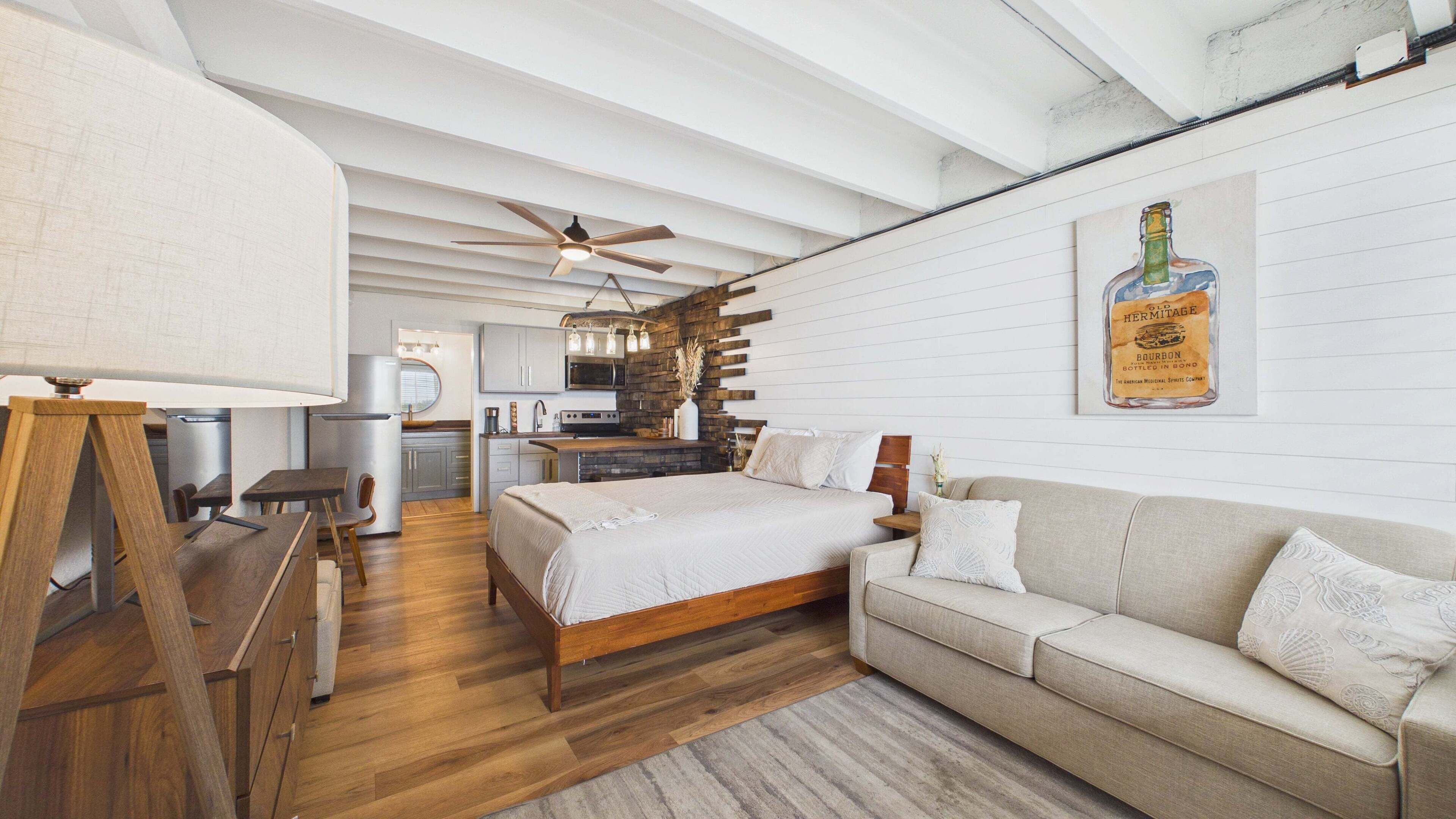 A modern bedroom with a wooden bed, a sofa, and a dining area with a kitchen in the background.