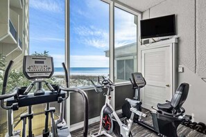 Ein Heimfitnessraum mit Trainingsgeräten, einem Fernseher und Blick auf den Strand.