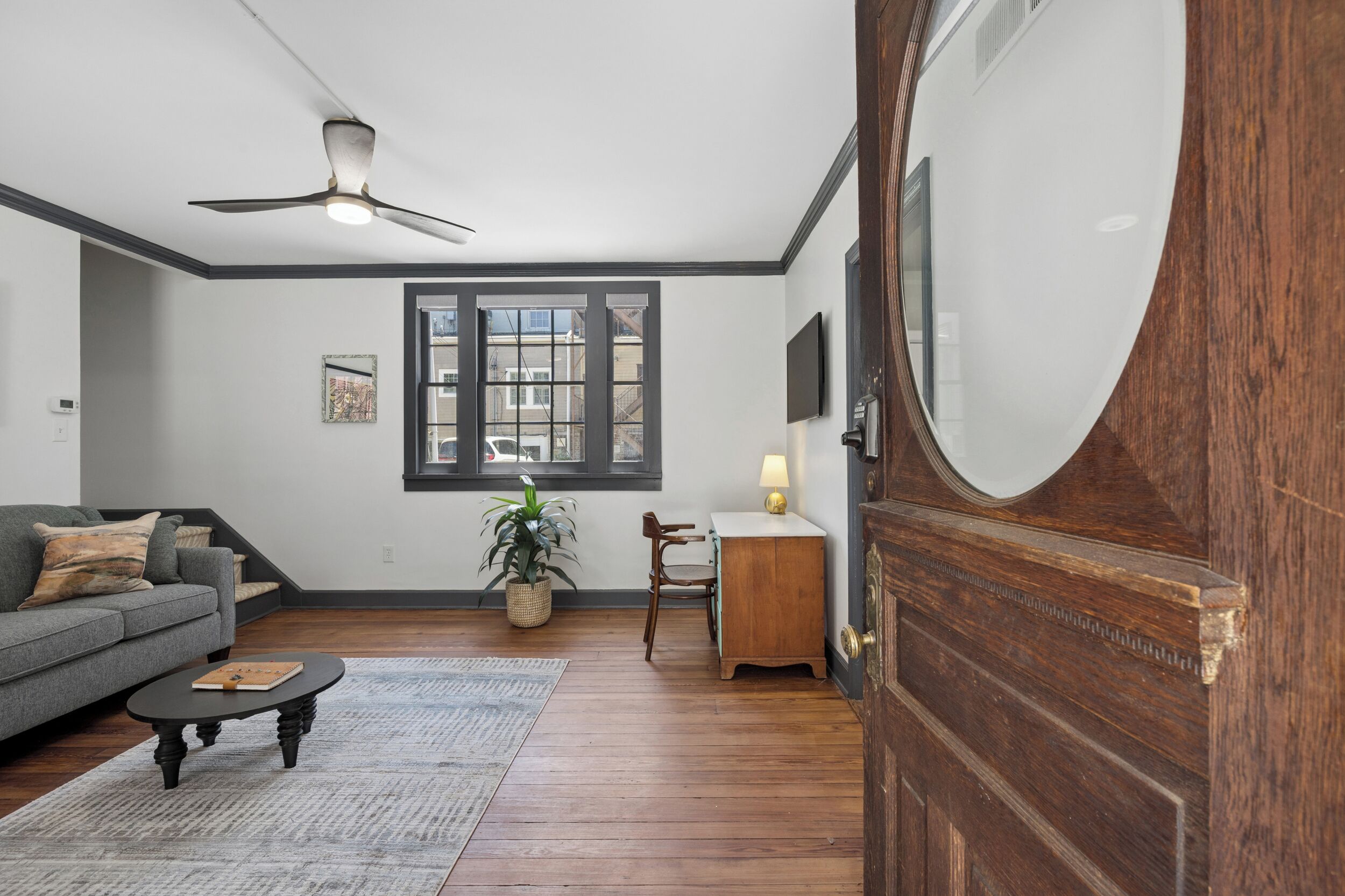 Quarto com ventilador de teto, sofá, mesa, cadeira, luminária, planta em vaso e porta de madeira.
