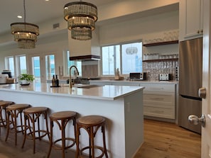 Una cocina moderna con una isla grande, taburetes de bar, un refrigerador de acero inoxidable y una araña.