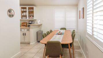 A dining area with a wooden table, green chairs, a kitchen with white cabinets, and a window with blinds.