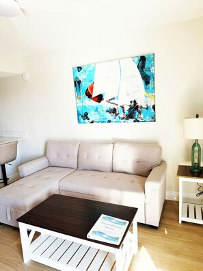 Un soggiorno con un divano angolare beige, un tavolino in legno, un quadro astratto appeso al muro, un piccolo tavolino laterale con una lampada e un cartello sul tavolino.
