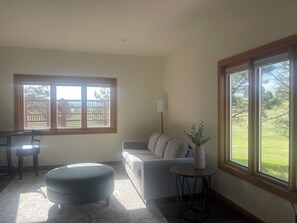 Ein Wohnzimmer mit einer Couch, einem Hocker und einem kleinen Tisch mit einer Vase. Zwei Fenster bieten Blick auf einen umzäunten Außenbereich.