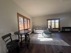 Un salón con sofá, reposapiés, mesa de comedor, sillas y ventanas grandes con vistas.