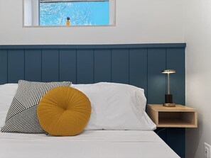 A bed with white and striped pillows, a mustard-colored round pillow, and a bedside table with a lamp.