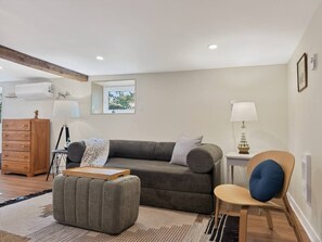 A modern living room with a grey sofa, a wooden side table with a lamp, a wooden chair with a blue cushion, and a wooden cabinet.