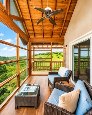 Ein Holzbalkon mit Korbmöbeln, einem Deckenventilator und großen Fenstern mit Blick auf viel Grün.
