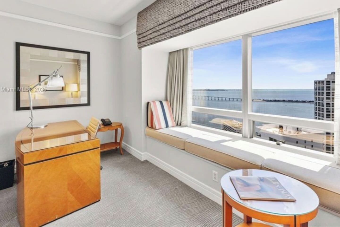 A hotel room with a large window, a desk, a chair, a small table, and a bed.