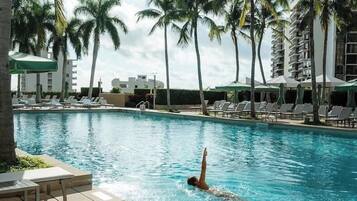 A swimming pool with a person swimming, surrounded by palm trees and lounge chairs.