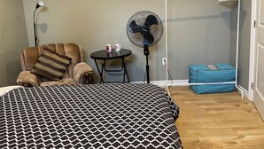 Um quarto com uma colcha estampada, uma cadeira com almofada listrada, uma mesinha com duas canecas, um ventilador e um móvel de armazenamento azul.