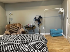 A bedroom with a patterned bedspread, a chair with a striped pillow, a small table with two mugs, a standing fan, and a blue storage unit.