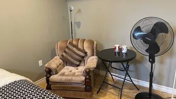 A room with a tufted armchair, a small round table with two mugs, a standing fan, and a bed with a patterned bedspread.