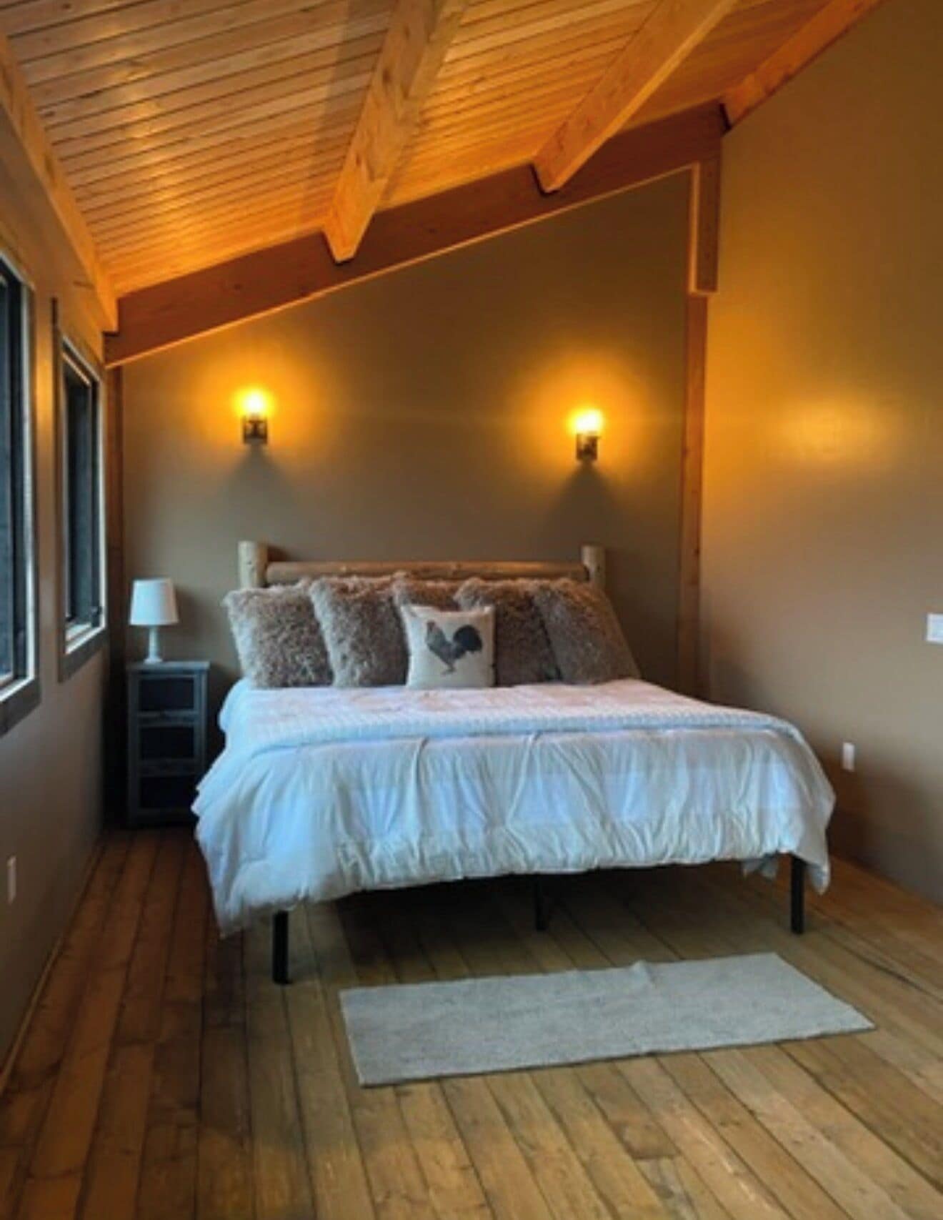 Ein Schlafzimmer mit Holzboden, zwei Wandleuchten, einem Bett mit weißer Bettdecke, einem Nachttisch mit Lampe und einem Fenster links.