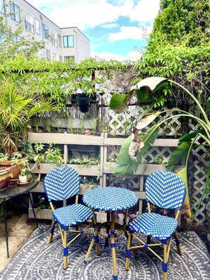 En terrasse med tre blå-hvide skakternede stole, en dekorativ måtte og flere krukker med planter.