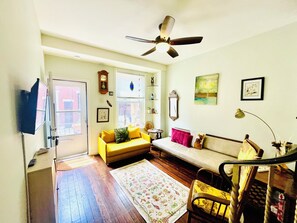 Ein Wohnzimmer mit Sofa, Sessel, Fernseher, Deckenventilator und Fenster.