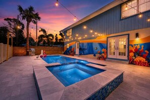 A swimming pool with a tiled deck, a mural on the building, and string lights.