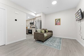 A modern living room with a sofa, a kitchen in the background, and a mounted TV. - The Nook - 20 mins from airport | Games | Coffee (Edmonton)
