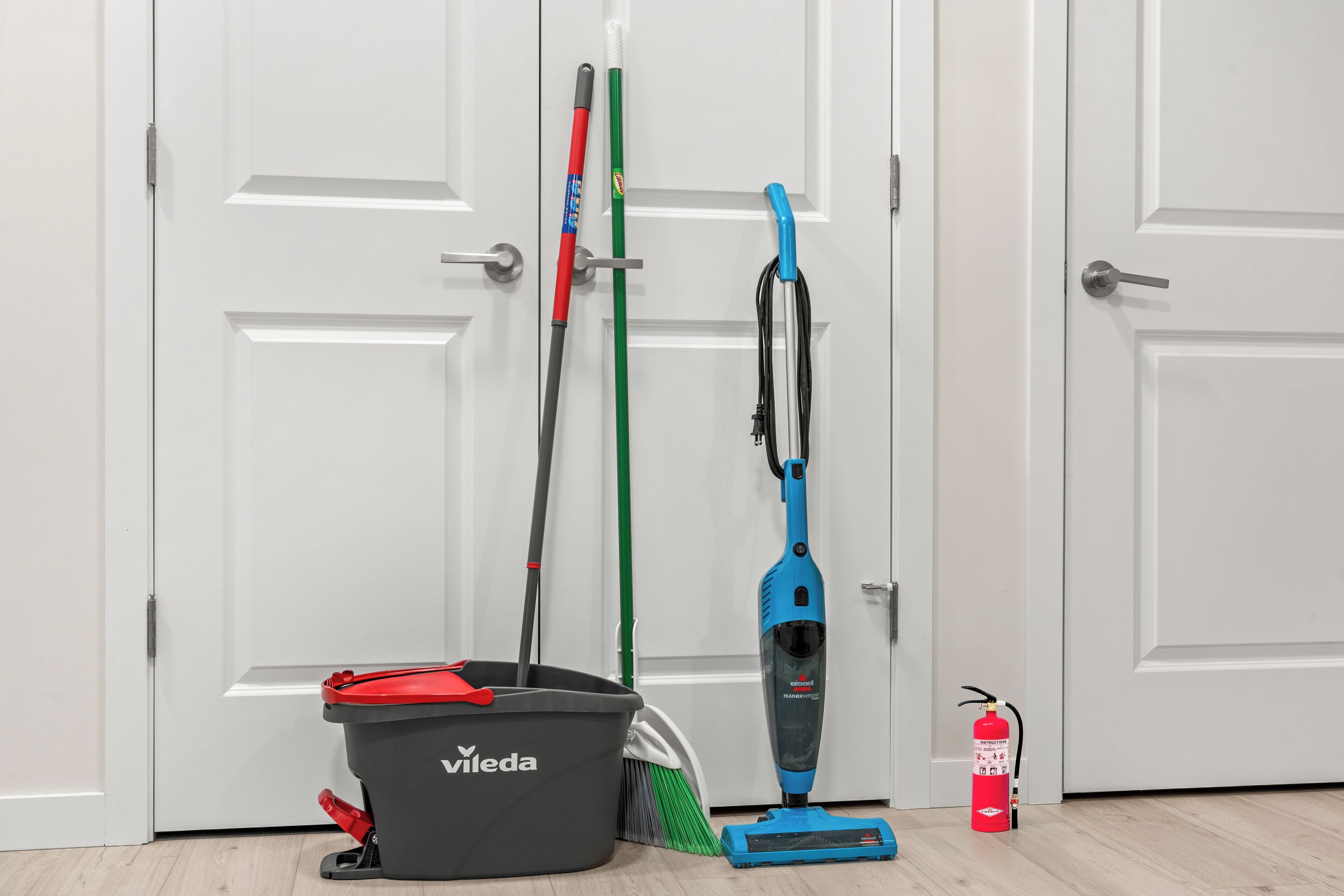 A room with a white door, a mop, a broom, a vacuum cleaner, a bucket, and a fire extinguisher.