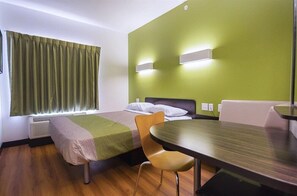 Hotellihuone, jossa on sänky, pöytä, tuoli, ikkuna verhoilla ja seinään kiinnitetyt valaisimet.