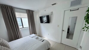 Un dormitorio con cama, televisor, puerta y ventana con cortinas.