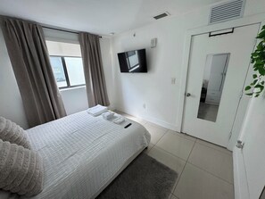 Ein Schlafzimmer mit Bett, Fernseher, Tür und einem Fenster mit Vorhängen.