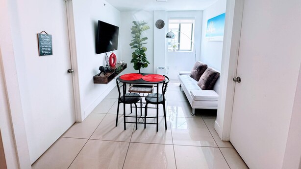 Una sala de estar moderna con un sofá blanco, una mesa de comedor con sillas, un televisor montado en la pared y un ventanal grande.