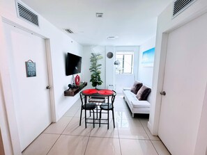 Ein modernes Wohnzimmer mit einem weißen Sofa, einem Esstisch mit Stühlen, einem an der Wand befestigten Fernseher und einem großen Fenster.