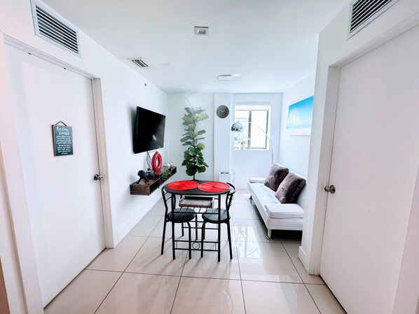 Un salon moderne avec un canapé blanc, une table à manger avec des chaises, un téléviseur fixé au mur et une grande fenêtre.