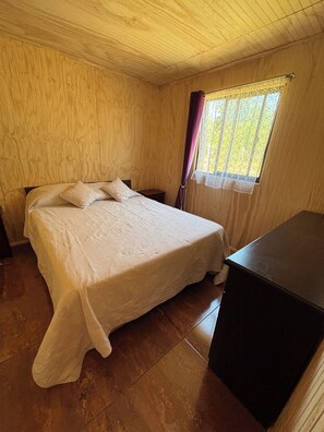 Ein Zimmer mit Holzboden, einem Bett, einem Nachttisch, einem Fenster mit Vorhängen und einer Kommode.