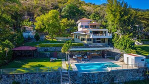 A multi-story house with a swimming pool, surrounded by lush greenery and hills.
