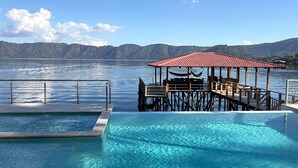 A swimming pool with a wooden deck and a covered seating area, surrounded by a clear blue sky and a calm lake.