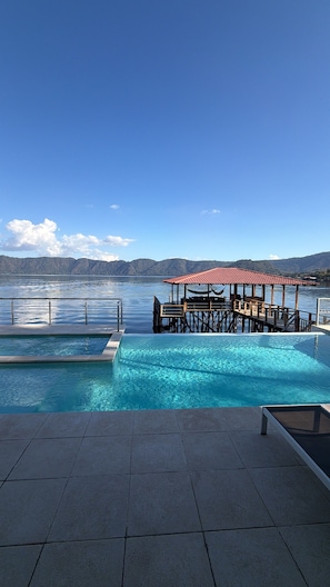Uma piscina com deck de madeira e área de estar coberta, cercada por um céu azul límpido e um lago calmo.