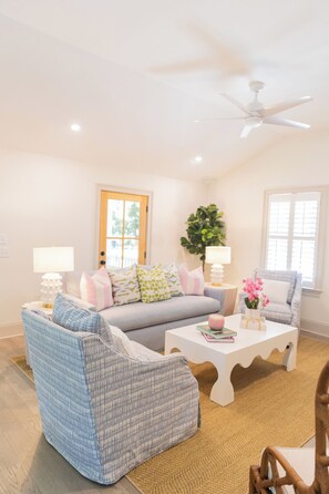 A living room with a sofa, two armchairs, a coffee table, a ceiling fan, a potted plant, and a door. - The Lettie | Newly updated Coastal Cottage (St Simons Island)