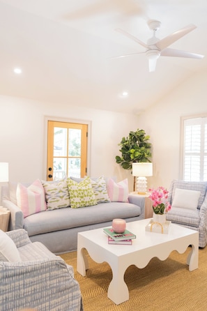 A living room with a white ceiling fan, a sofa with patterned cushions, a white coffee table, and a side table with a lamp. - The Lettie | Newly updated Coastal Cottage (St Simons Island)