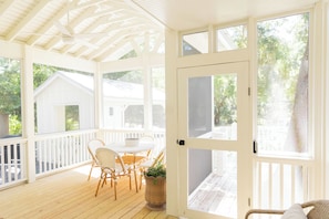 Un porche couvert avec une table et des chaises, une porte blanche et une vue sur une maison.