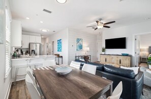 Eine moderne Küche mit weißen Schränken, ein Essbereich mit einem Holztisch und Stühlen, ein schwarzes Ledersofa und ein Deckenventilator.