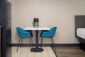 Ett litet rum med ett vitt bord, två blå stolar, en kaffebryggare, en säng och ett kylskåp.