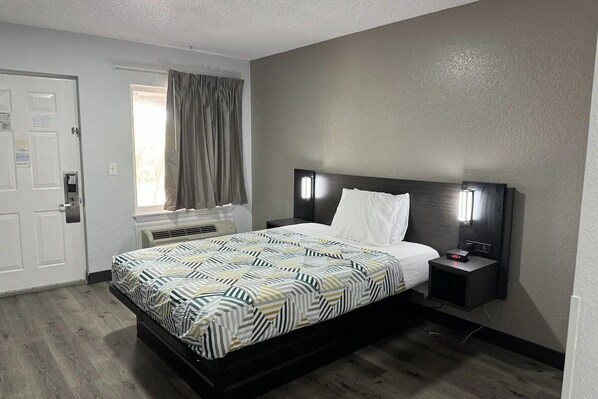 A hotel room with a bed, bedside table, nightstand, window with curtains, and a door.