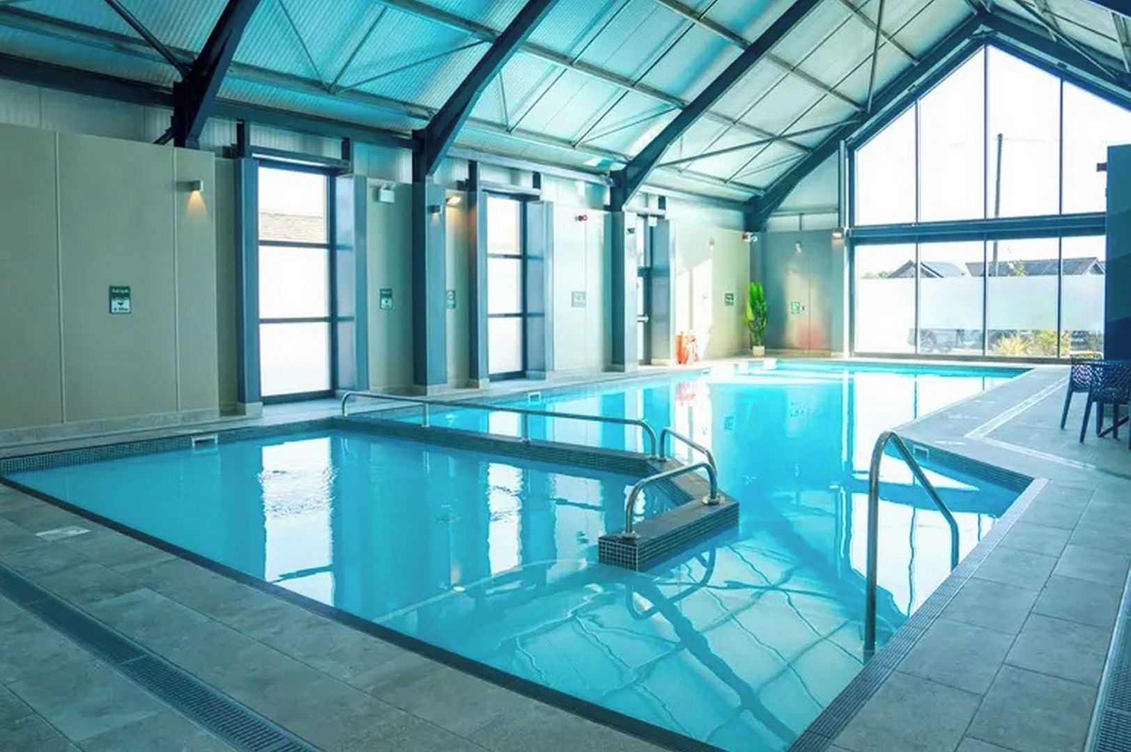 An indoor swimming pool with clear glass walls, a tiled floor, and a metal roof structure.
