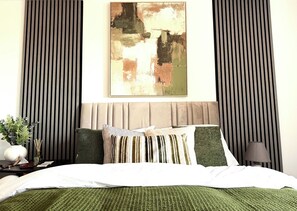 Ein Schlafzimmer mit einem Bett, Kissen, einem Nachttisch mit Lampe sowie einem Wandbild im abstrakten Stil.