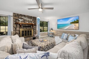 Ein Wohnzimmer mit Kamin aus Stein, Deckenventilator, großem Fernseher mit Strandmotiv und einer gemütlichen Sofagarnitur.