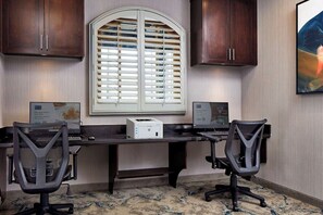 Ein Zimmer mit Schreibtisch, zwei Bürostühlen, einem Drucker, einem Fenster mit Jalousien und einem Schrank.