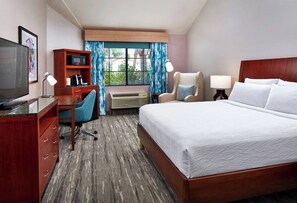 Quarto de hotel com cama, escrivaninha, cadeira, televisão, minibar e janela com cortinas.